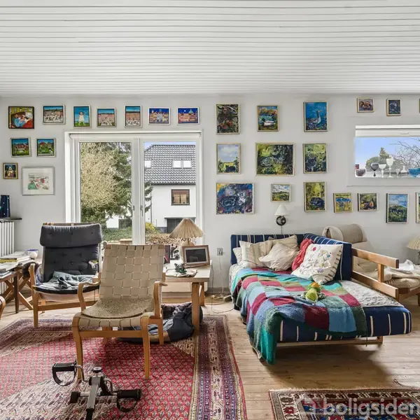 Stue med farverig sofa dækket af tæppe, omgivet af stole og borde. Væggene er dekorerede med mange malerier. Vinduer giver udsigt til en have og hus. Stor gulvtæppe pryder gulvet.