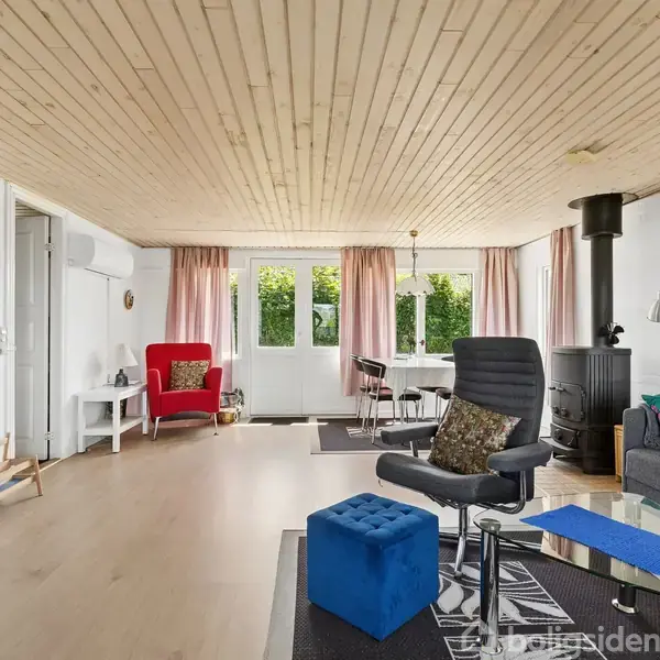 Stue med moderne møbler; lænestol, sofa, og spisebord, placeret ved vinduer. Sort brændeovn står i hjørnet. Lyse trægulve og loft skaber en lys og rummelig atmosfære.