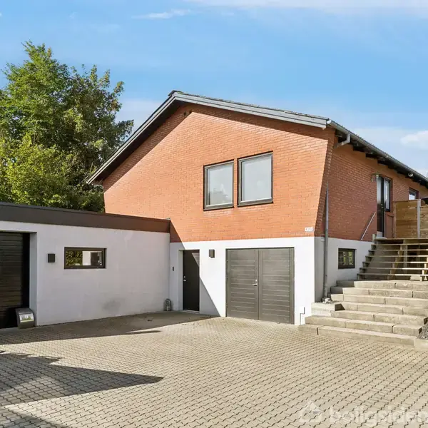 Rødt murstenshus med trappe, stiller i et solrigt område. Garage med sorte døre ved siden af, flisebelagt indkørsel og grønne buske omgiver pladsen.