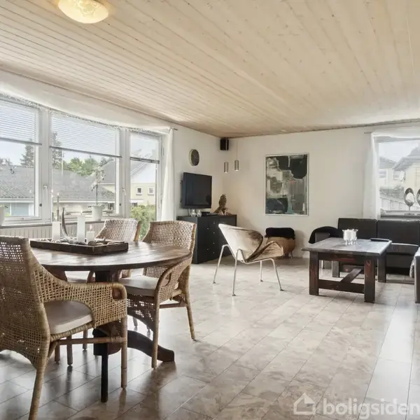 Stue med spisebord og stole af træ placeret på stenfliser, sofa og sofabord bagerst til højre. Store vinduer slipper lys ind, udsigt til et rødt vinglas.