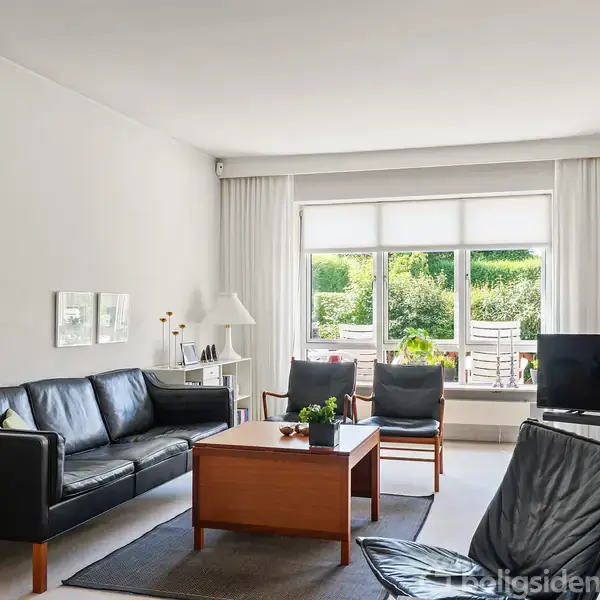 En sort sofa står i en stue med lyse vægge. Foran sofaen er et træbord. Store vinduer i baggrunden lukker naturligt lys ind.