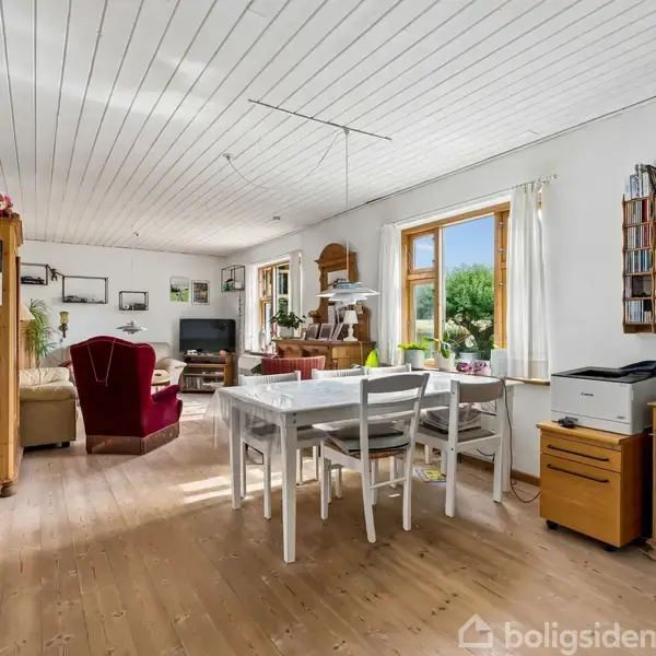 Spisestue med hvidt bord og stole er placeret ved et stort vindue, der slipper naturligt lys ind. I baggrunden er en sofa, reoler med cd'er og planter.