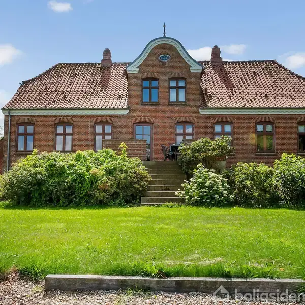 Rødt murstenshus med symmetrisk facade og tegltag, omgivet af grønne buske og velplejet græsplæne i solrigt vejr.