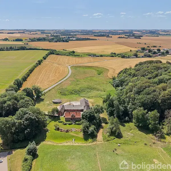 Hus omgivet af skov, beliggende i et landligt område med marker i baggrunden under en klar blå himmel.