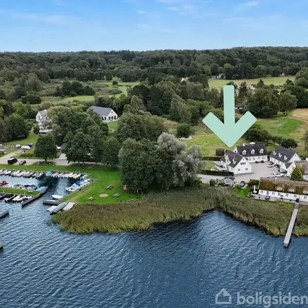 Hvid bygning med stråtag ligger ved vandkanten, markeret med en grøn pil. Omkringliggende skov og marina med både danner en naturskøn kontekst.