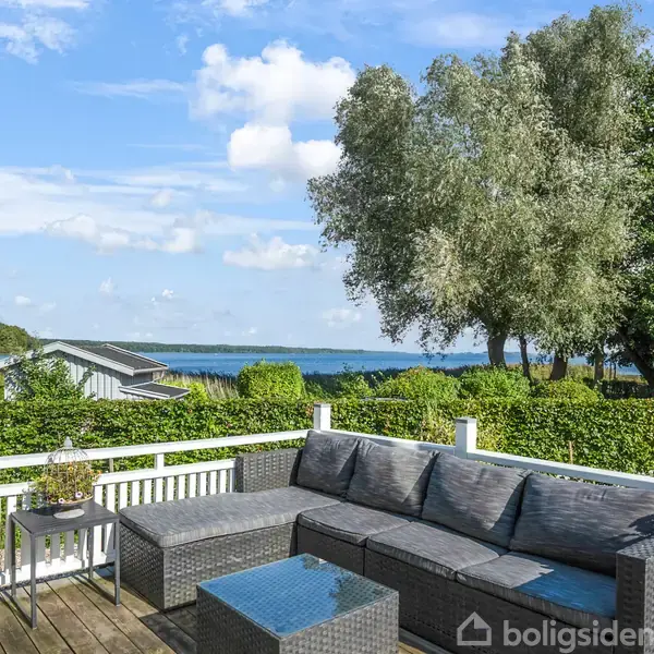 Rundt spisebord med træstole står ved franske døre, der åbner til terrasse med sofa, omgivet af grønne træer. Hvidt interiør med trægulve. Køkkenø til venstre.