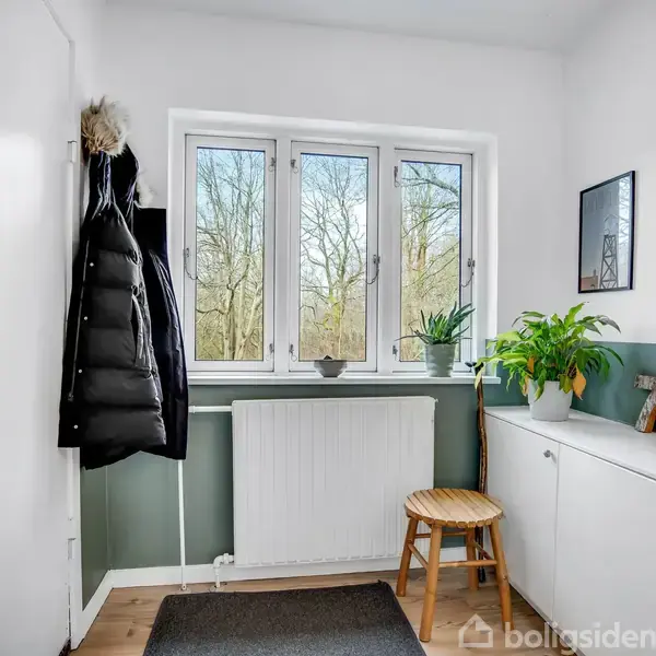 En grå hjørnesofa står i stuen, omgivet af puder, et sofabord og en stående lampe. Store vinduer slipper naturligt lys ind, og en farverig plakat pryder væggen.