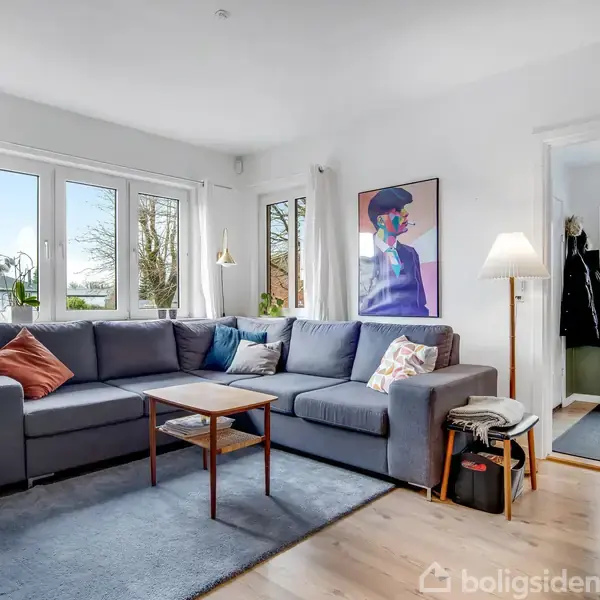 Stue med en grå sofa omkranset af puder, et lille sofabord, og en fladskærm på en bænk. Brændeovn med brænde ved siden af og store vinduer med udsigt.