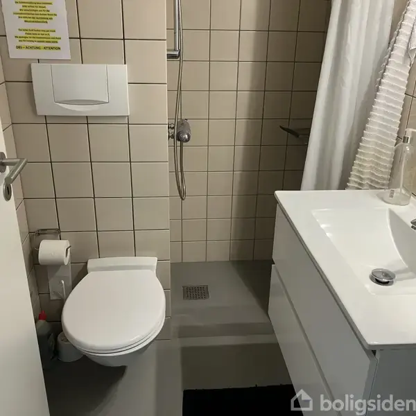 Toilet og håndvask står på gråt flisegulv. Bruser med gardin til højre. Væggene er flisebeklædt. Tekst på væg: “W.C. UDEN AFKALKNING…”. Miljøet er et moderne badeværelse.