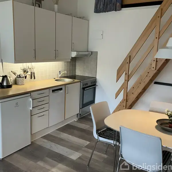 Et køkken med hvidt skabsmøblement, komfur og lille køleskab, ledsaget af et rundt spisebord med fire stole. Trætrappe fører opad mod en højere etage.