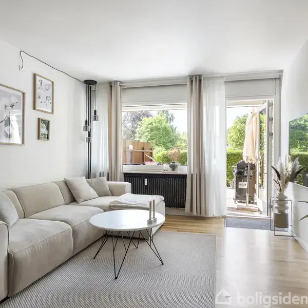Stue med beige sofa ved et rundt sofabord. Store vinduer og åben dør fører ud til terrasse med grønne planter. Vægkunst og lampe tilføjer indretning.