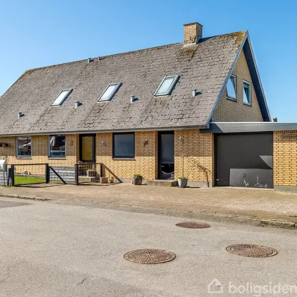 Hus udført i gule mursten med en mørk tagflade og flere tagvinduer. Det står i et boligområde med garage, fortov og vej foran.
