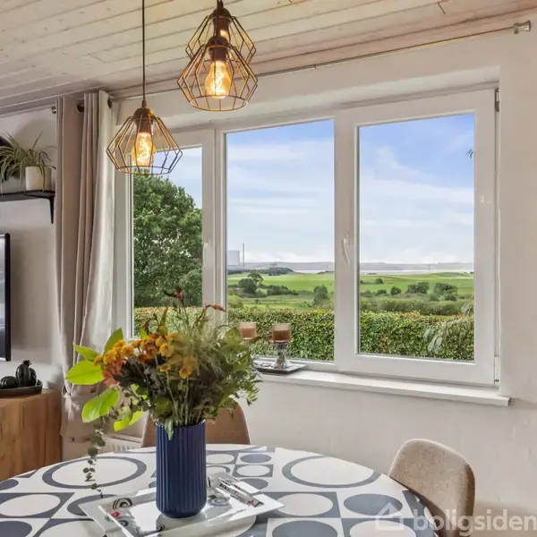 Et rundt bord med en vase af blomster står i en lys stue med store vinduer, der viser en grøn udsigt. En plakat med teksten "Ruth Orkin" hænger på væggen.