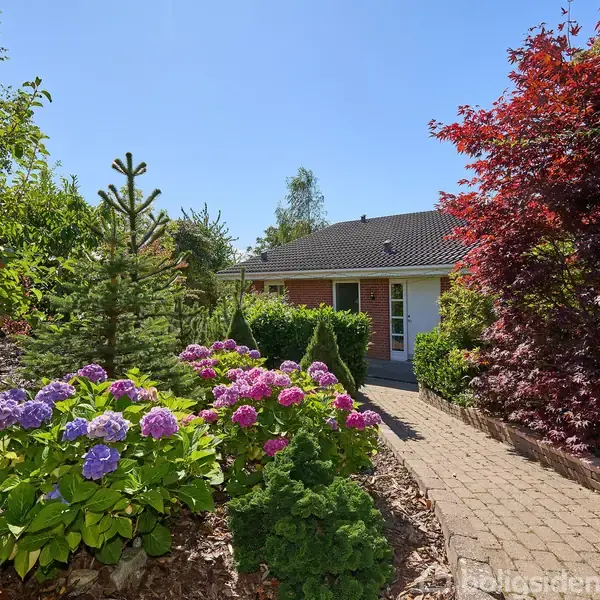 Et hus med rød murstensfacade omgivet af en farverig have. Foran huset er der en brolagt sti, omgivet af grønne og blomstrende planter under en klar blå himmel.