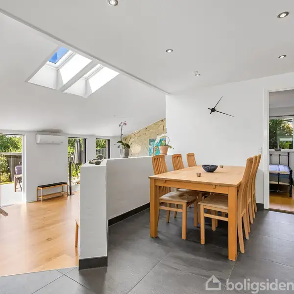 Spisebord med seks stole står i en moderne, lys stue. Ovenlys og vinduer lukker dagslys ind. Trappe fører til højre, og et åbent soveværelse ses i baggrunden.