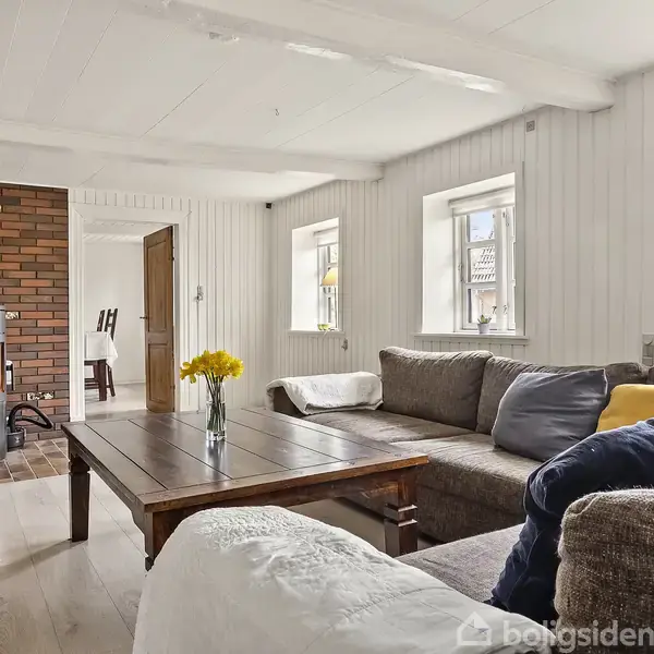Brændeovn varmer i stue med lyse vægge. Brun sofa med puder står ved træbord med gule blomster i vase. Vinduer lukker lys ind.