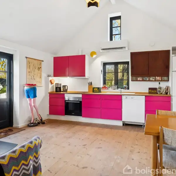 Et moderne køkken har pink skabe, et brændeovn med ild, og et bord med stole. Vinduer lukker lys ind. En gulvlampeskulptur af en mand i blå shorts står i hjørnet.