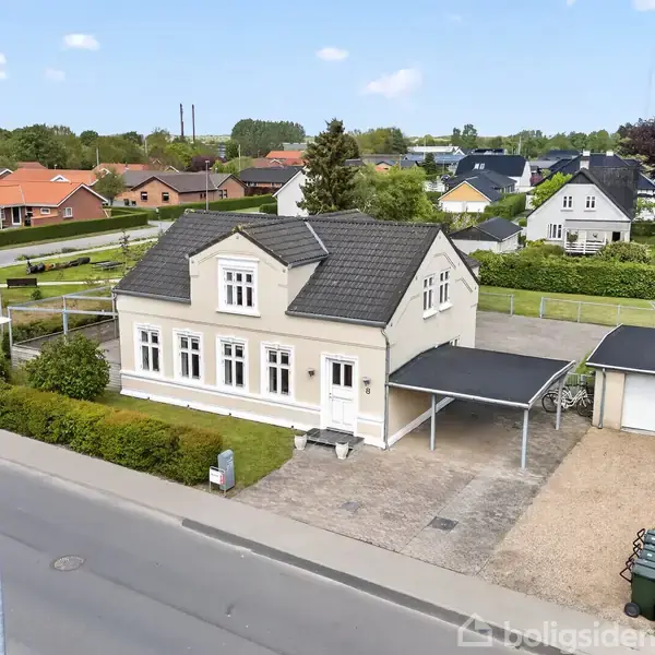 Et to-etagers hvidt hus med sort tag står ved en vej. Der er en overdækket carport og en have bagved. Omkringliggende boliger og træer ses i baggrunden.