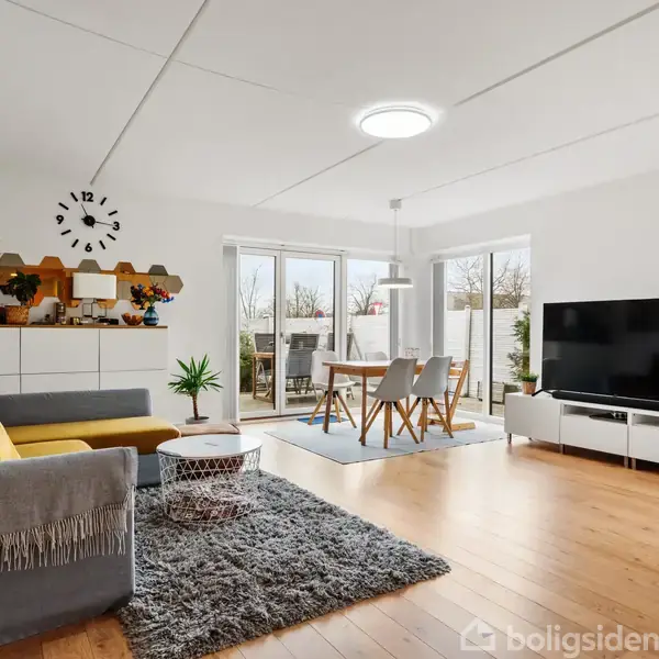 En moderne stue med en grå sofa, et lille bord, og et gulvtæppe. Et stort TV står på en hvid reol. Glastæppeborde fører ud til terrassen. En vægdekoration inkluderer et ur.