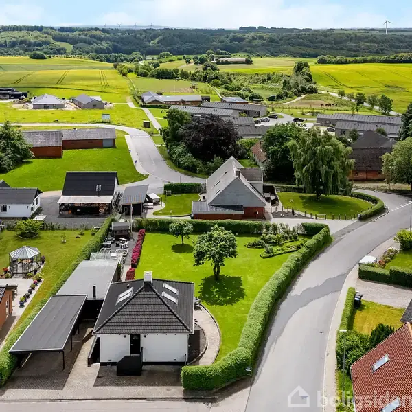 Række af villaer med sort og hvidt tag, omgivet af velplejede haver; en bil på indkørslen nær en hævet vej. Grønne marker og bakker danner baggrunden. Ingen tekst til stede.