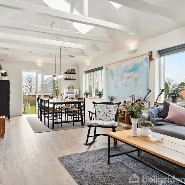 Stue med grå sofa og træbord foran et stort vindue. Spisebord og stole i baggrunden, omgivet af hvide vægge med verdenskort. Lys og moderne indretning.