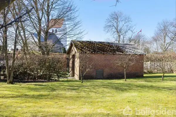 Et lille, rødt murstensskur står stille i en solrig have omgivet af træer. En kirkebygning med klokketårn ses i baggrunden. Ingen tekst er synlig.