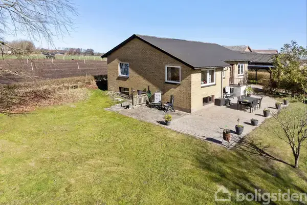 Et gulstenshus står på en åben græsplæne med en flisebelagt terrasse. Havemøbler står på terrassen, og i baggrunden ses dyrkede marker og spredte træer under en klar himmel.