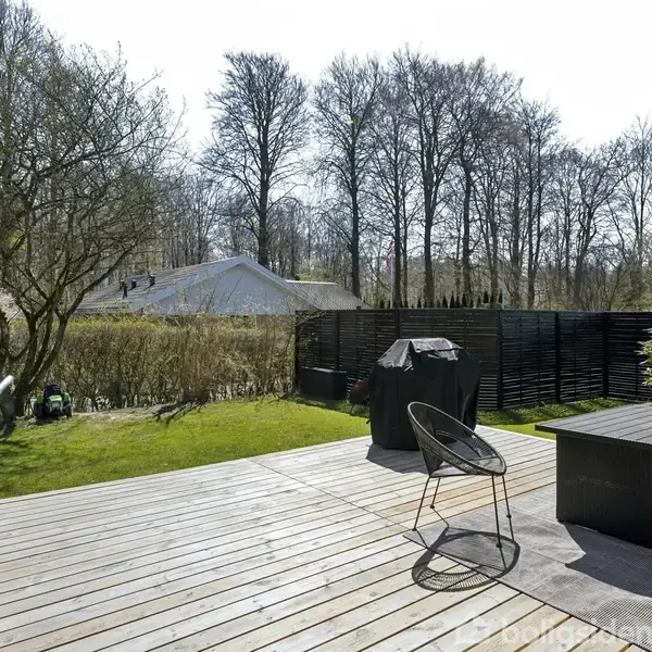 Terrassen har en trådstol og en grill, omkranset af græs og træer i baggrunden. Hækken skaber en grøn ramme, mens høj sort hegn giver afskærmning.