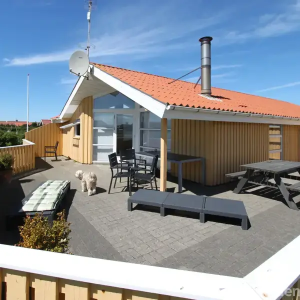 Hus med terrasse: Et træhus med rødt tag har en stor, møbleret terrasse med liggestole og et bord/bænkesæt. En hund står på fliserne. Omgivet af grønt landskab under blå himmel.