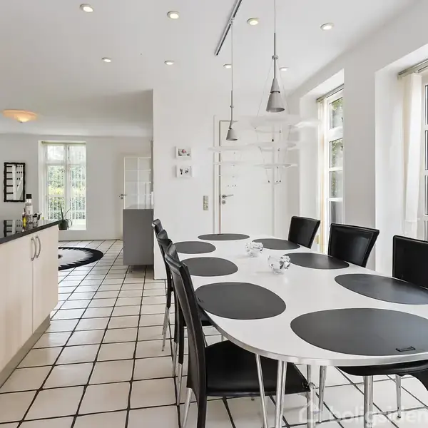 Lang hvidt spisebord med sorte dækkeservietter og sorte stole står i et moderne køkken. Køkkenet har lyse gulvfliser, hvide skabe og store vinduer med udsigt til en grøn have.