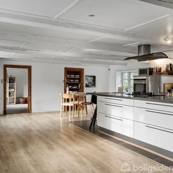 Køkkenø med skuffer står centralt, omgivet af spisebord og hylder, i et lyst rum med træloft og gulv. Flere vinduer giver naturligt lys.