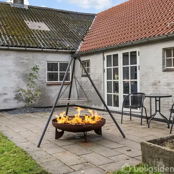 Bål i en rund ildskål brænder klart under en grillrist på en gårdsplads med stole og bord. Omkranset af hvidt murstenshus med rødt og gråt tag.