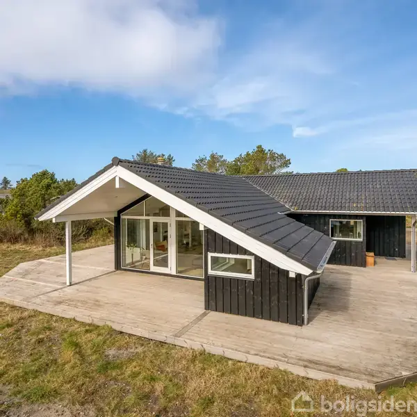 Sort træhus med store vinduer står på flisebelagt terrasse. Omgivet af græsplæne og buske under en blå himmel.