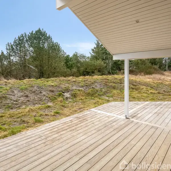 Træterrasse under overdækket del i et naturområde. Omkranset af græsklædte klitter og buske, mens himlen ses i baggrunden. Der er ingen tekst i billedet.