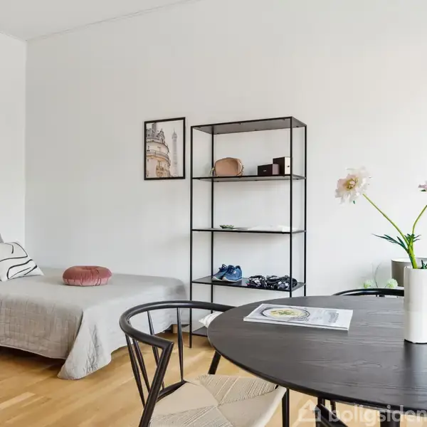 En enkeltseng med gråt sengetæppe står ved siden af en metalreol mod en hvid væg; et rundt bord med en vase og magasin står foran med træstol.