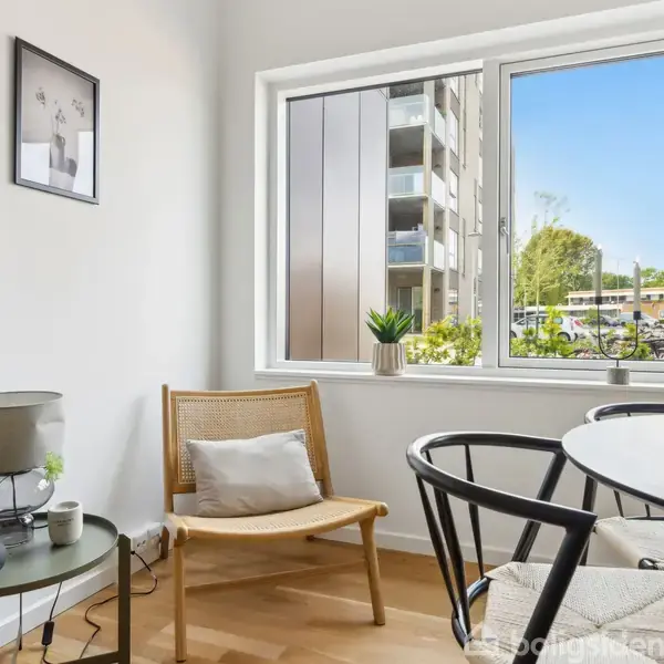 Stol og bord står i et lyst rum med hvidmalede vægge og trægulv. En stor, firkantet vindue viser en udsigt til en moderne bygning og nogle grønne træer udenfor. En lampe og planter er på bordet.