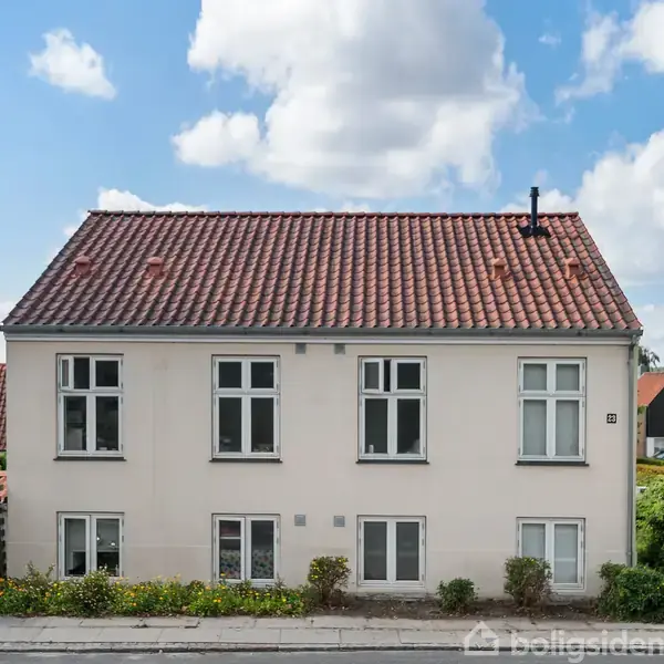 Et hvidt hus med rødt tegltag og flere vinduer står langs en vej. Buske pryder husets facade, og en blå himmel med skyer ses i baggrunden.