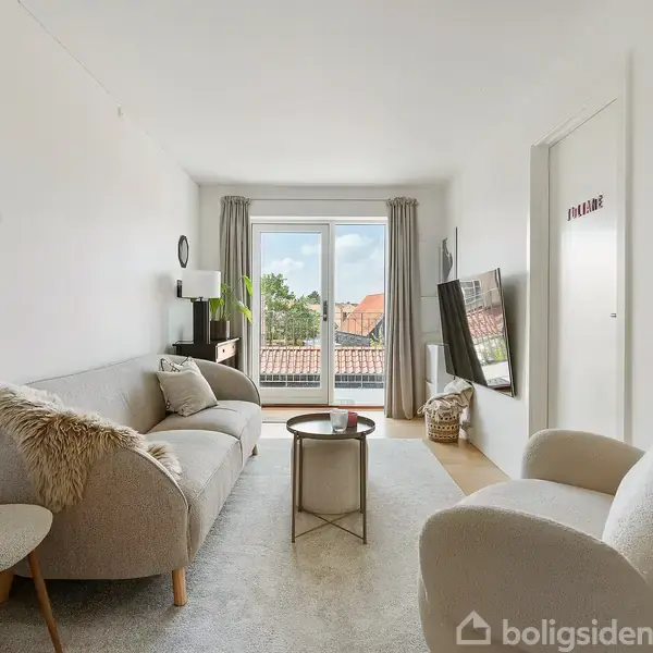 En lys stue med sofa og to stole, en vægmonteret TV-skærm til højre og glasdøre i baggrunden. Døren har teksten "WELCOME". Rummet er indrettet med stilfulde, neutrale møbler.