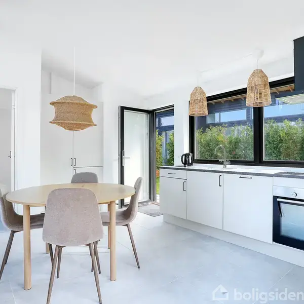 Spisebord med fire stole står i et lyst køkken. Køkkenet har hvide skabe, indbygget ovn og store vinduer, der giver udsigt til grønne buske udenfor.