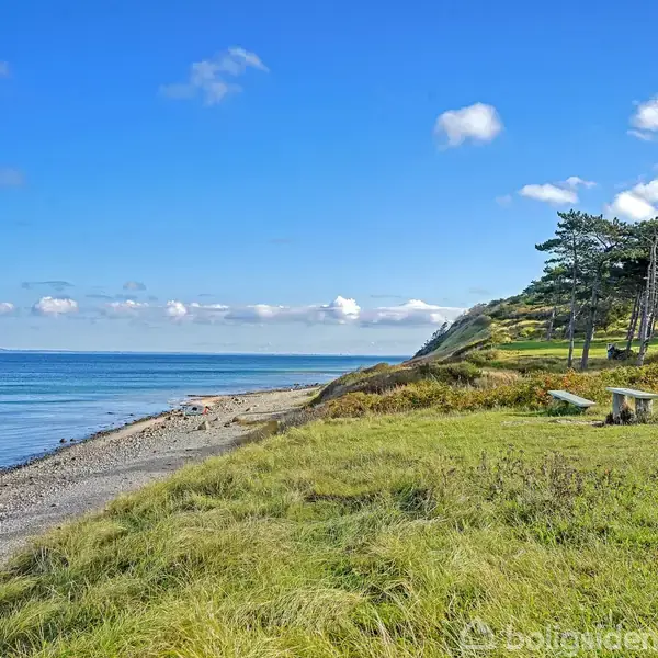 Strand med græsbevoksede bakker og stenet kyst. Træer står på en bakke til højre. Blå himmel med få skyer hænger over det stille hav.