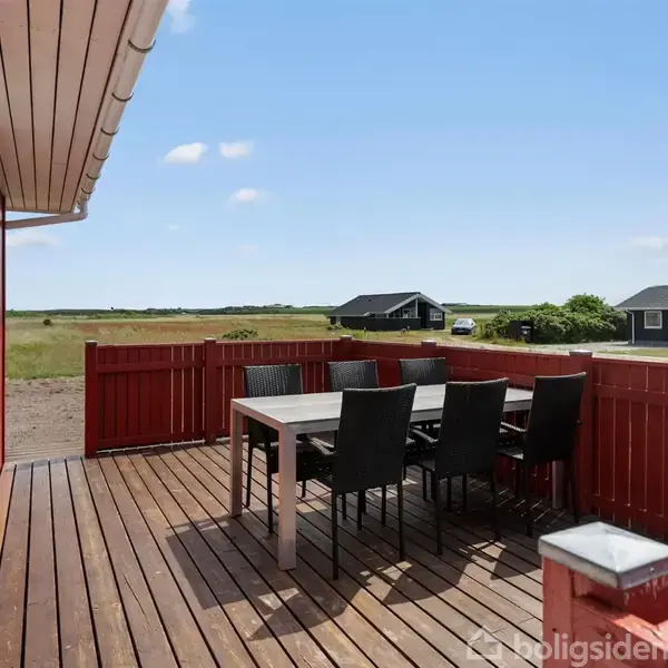 En træterrasse med et bord og seks sorte stole står foran et rødt hus. I baggrunden ses en åben mark med enkelte huse ved horisonten under en blå himmel.