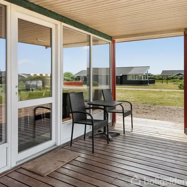 Terrasse med to sorte havestole og et bord står foran et hus med store glasdøre. Området omkring er dækket af græsplæner og andre huse kan ses i baggrunden.