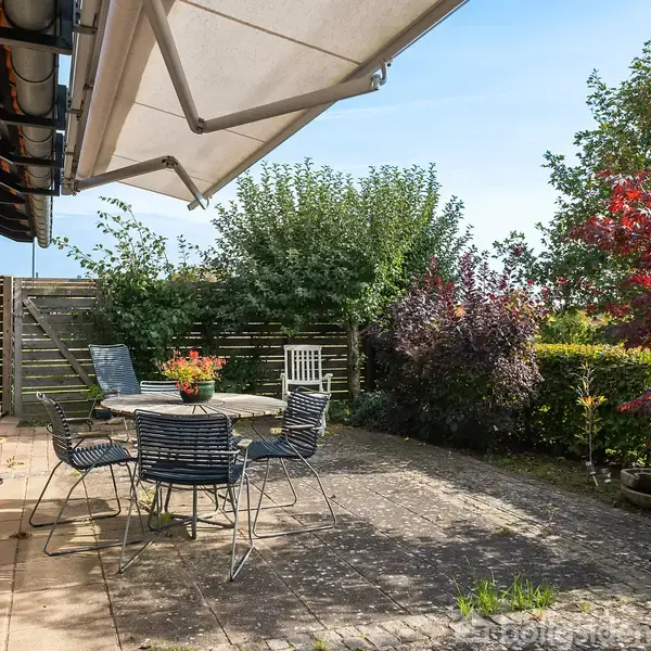 En terrasse med et rundt bord og fire stole står stille under en markise. Omgivet af en frodig have med forskellige buske og træer i baggrunden.