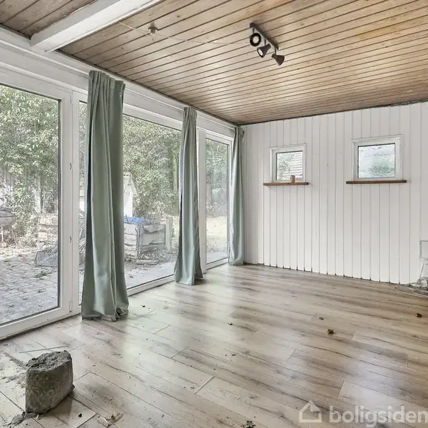 Stue med trægulv og træloft, store glasdøre med grønne gardiner, åben til terrasse. To små vinduer i hvidmalet panelvægt. Udenfor ses træer og hegn.
