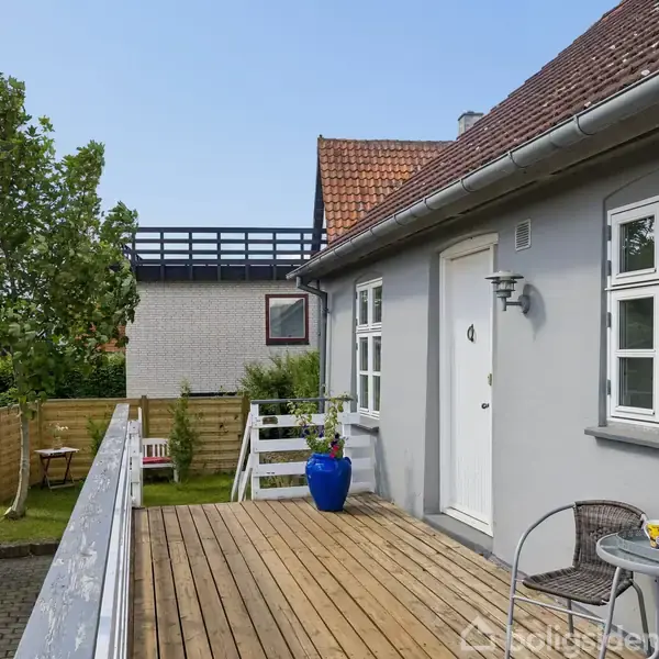 En træterrasse langs et hus har et cafébord med stole og blomster. En blå urtepotte står ved døren. Baggrunden indeholder et hegn og et træ.
