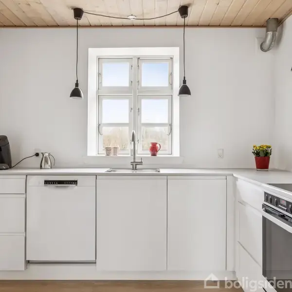 Hvidt køkken består af moderne skabe og apparater; en kaffemaskine og planter pryder bordet. Loftet er træ, og vinduet centralt lader lys trænge ind, mens to hængelamper hænger over bordpladen.