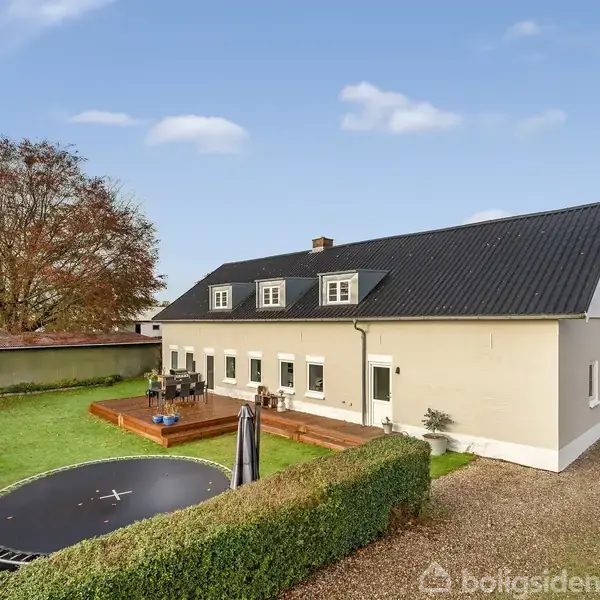 Et beige hus med sort tag har en træterrasse foran. I haven er en nedgravet trampolin omringet af hække. Det er solskinsvejr, og der er et stort træ i baggrunden.