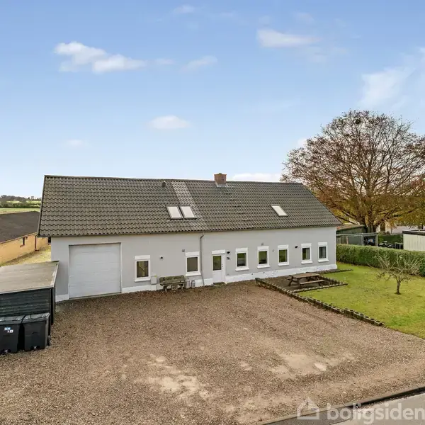 En hvid bygning med sadeltag står i et område med grusbelagt forplads og grønne græsarealer. Omkringliggende hække og træer omkranser huset, der har flere små vinduer.