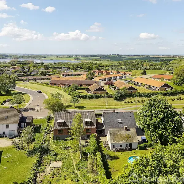 Huse med haver står i række, omgivet af frodige grønne områder og træer. I baggrunden ses flere bygninger samt åbne marker under en blå himmel med få skyer.