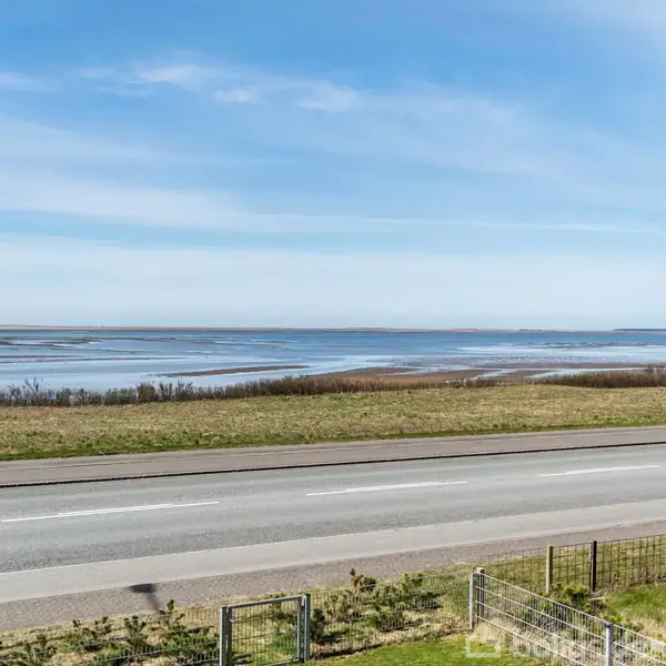 Vejen strækker sig langs en kystlinje, hvor havet møder horisonten under en klar blå himmel. Græsarealer og buske flankerer vejen, hvilket skaber en fredelig scene.